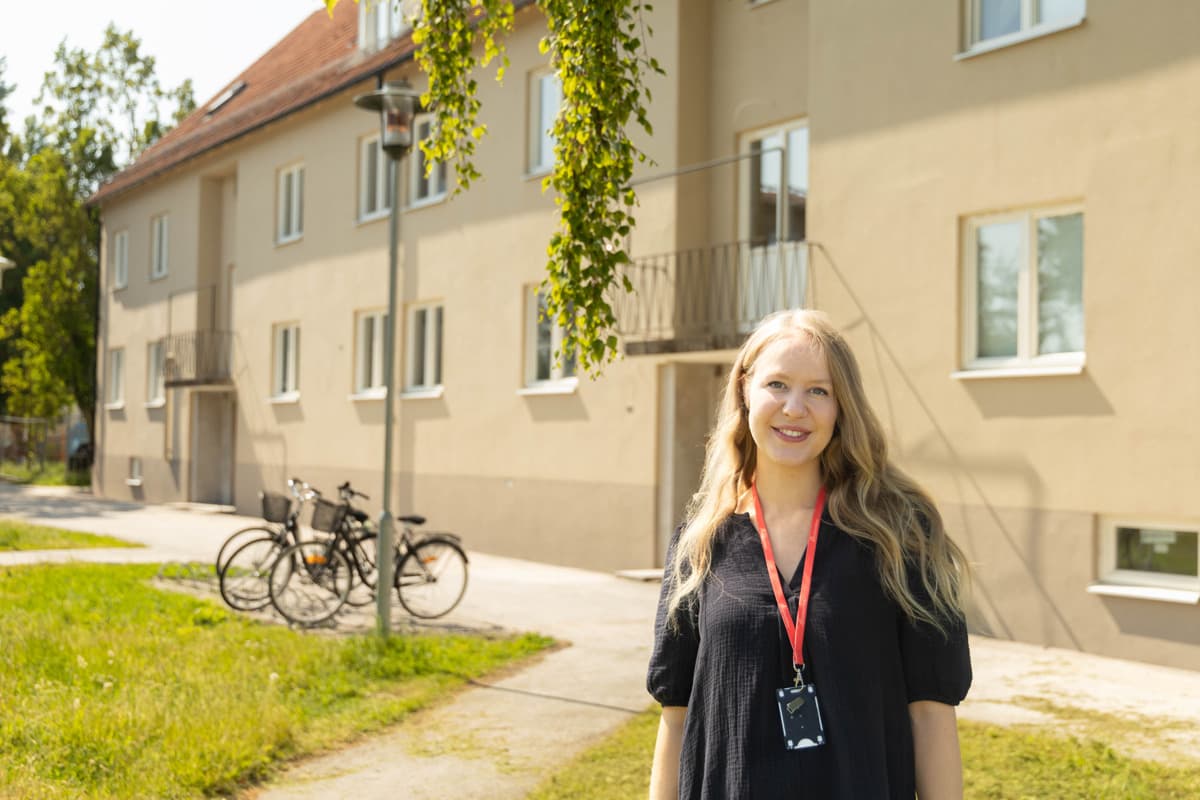 En kvinna med långt blont hår och svart tröja står framför ett gult bostadshus. Solen skiner.