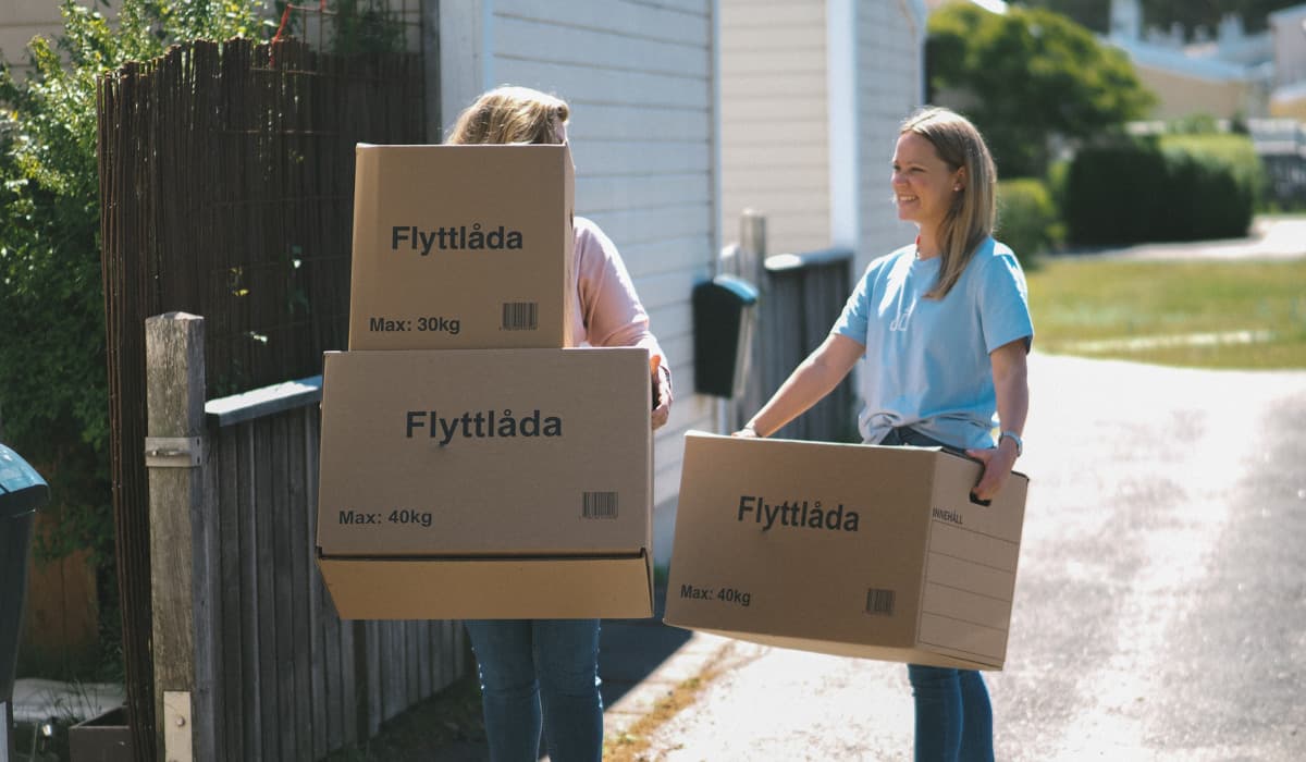 Två kvinnor bär på flyttkartonger. Den ena kvinnan har en rosa tröja och bär två kartonger, den andra kvinnan har en ljusblå t-shirt och bär en kartong. De är utomhus i ett bostadsområde.