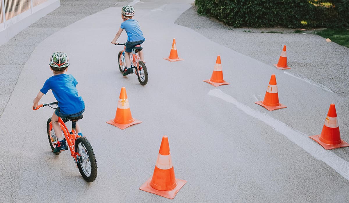 Två små barn i blå tröjor och färgglada cykelhjälmar cyklar genom organge koner.