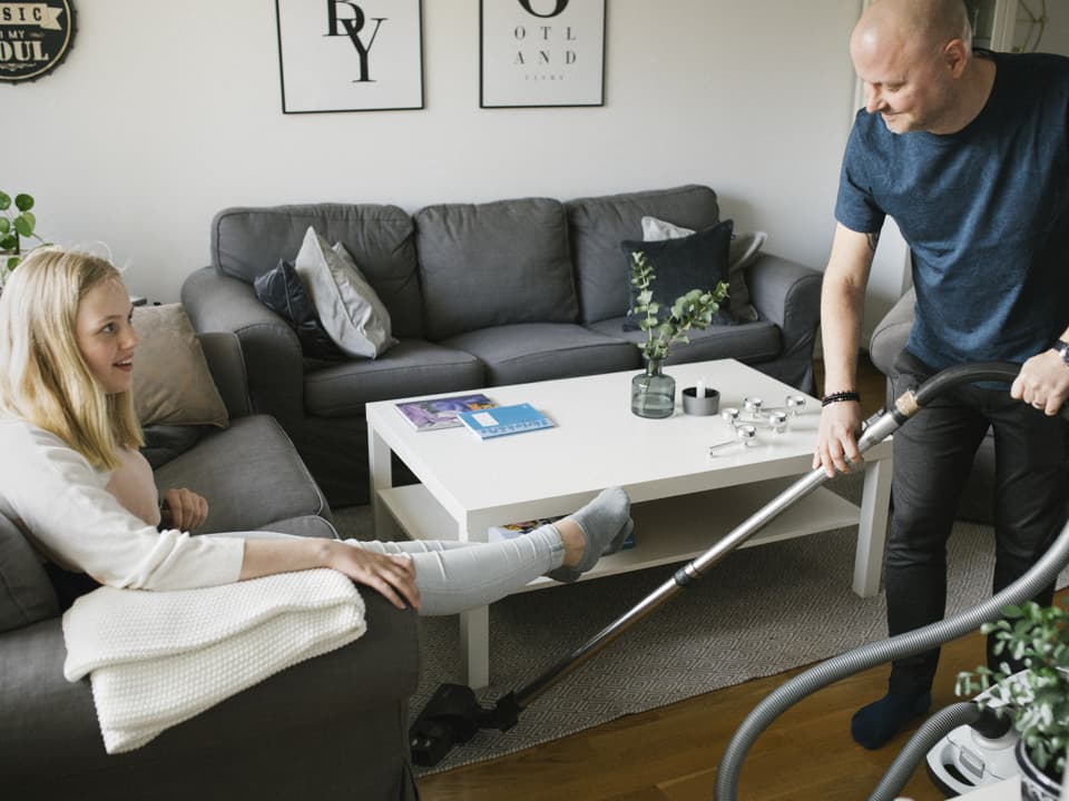 En man håller i en dammsugare och dammsuger under en grå soffa. På soffan sitter en tjej.