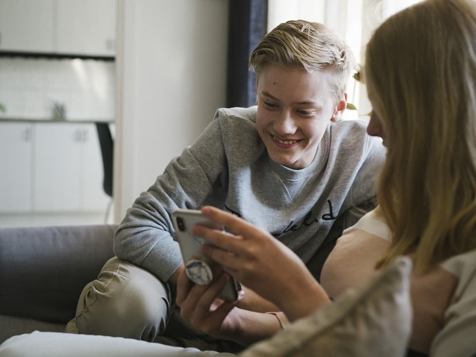 Två tonåringar, en tjej och en kille, sitter i en soffa i ett vardagsrum och kollar på en telefon.