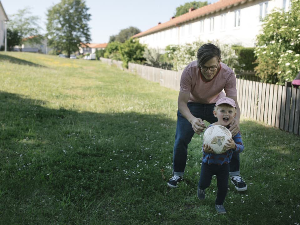 En pappa och ett barn leker på en grön gräsmatta. Barnet håller i en fotboll. I bakgrunden syns hus och ett staket.