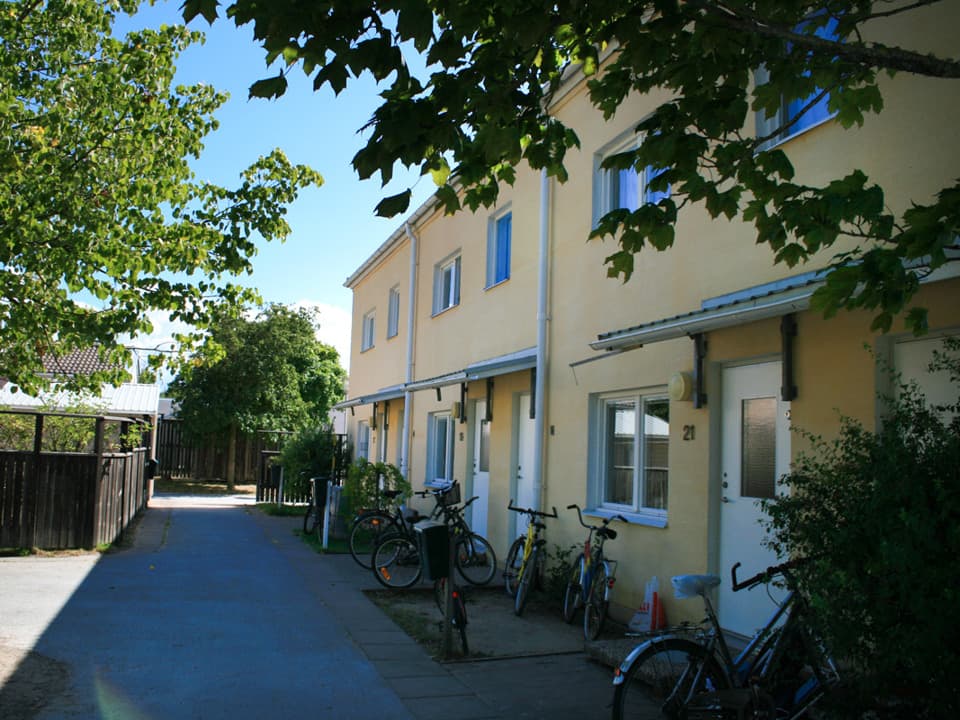 Ett gult tvåvåningshus med vita dörrar och fönsterkarmar. Framför huset står cyklar parkerade och det går en smal asfalterad cykel- och gångväg. Runtom huset är det gröna lövträ och himlen är blå.