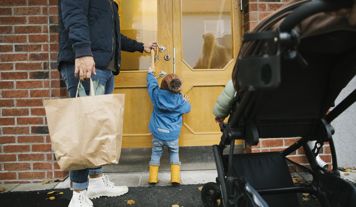 Ett barn sträcker sig för att nå handtaget till en ytterdörr. På ena sidan står en vuxen som håller i en kasse och hjälper barnet med dörren. På andra sidan står en barnvagn.