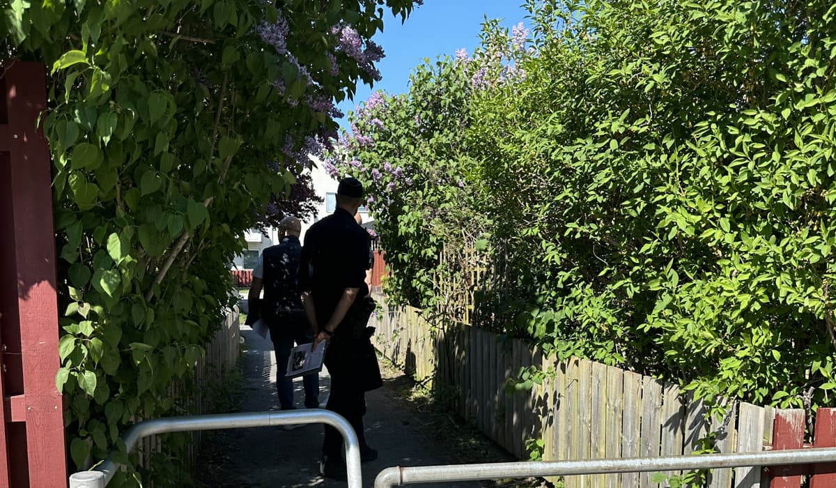 Polis och person från GotlandsHem tittar på en passage in till ett av GotlandsHems bostadsområden. Runt dem grönskar häckar och buskar. En stor lila syren blommar. Det är sommar och sol.