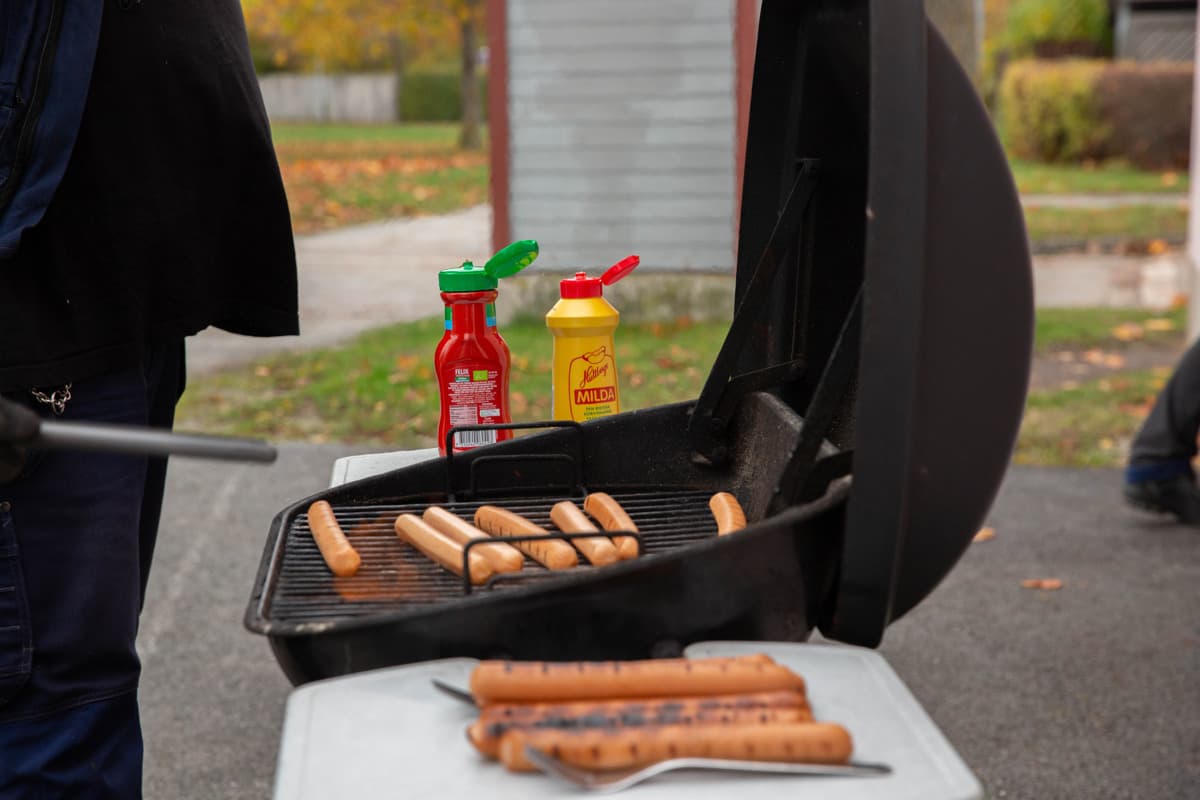 En grill står öppen, det ligger korvar som grillas i grillen. I förgrunden syns färdiggrillade korvar och i bakgrunden står en ketchupflaska och en senapsflaska. Det är städdag och korvgrillning.