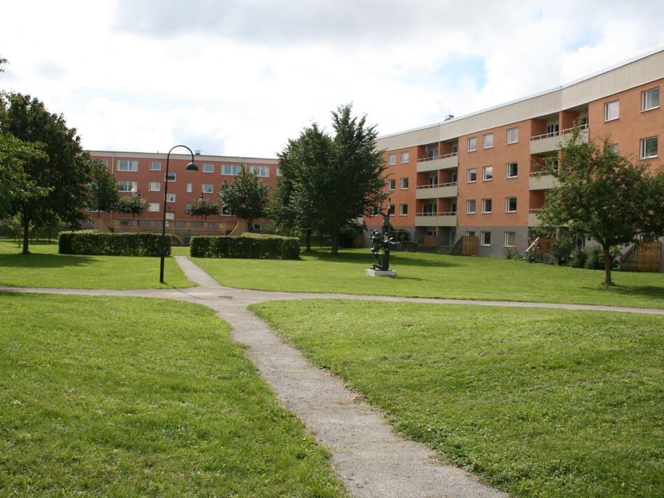 Två bostadshus, ett rött och ett orange. Framför dem är ett grönområde med buskar, träd och statyer. Genom gräset går smala stigar. 