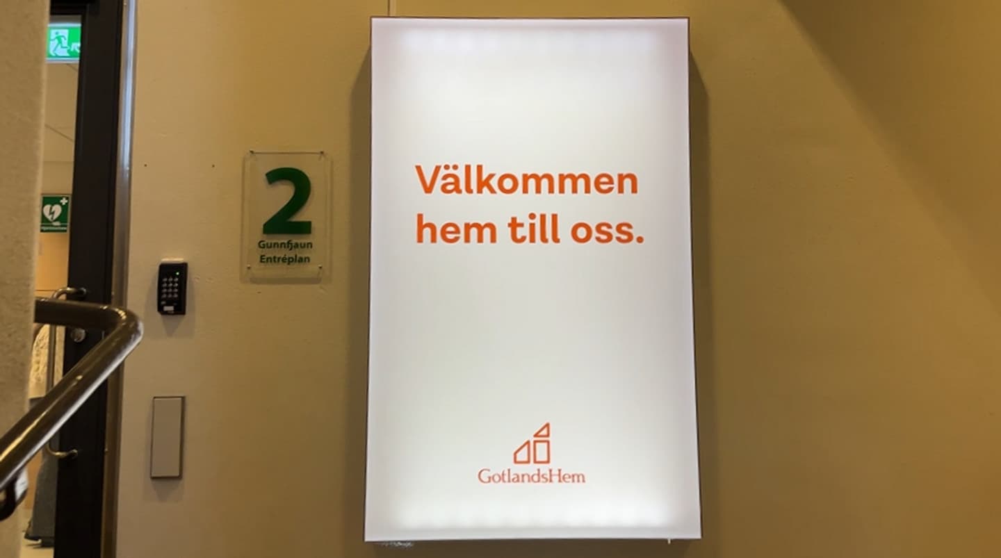 En ljusrosa skylt med röd text som står "Välkommen hem till oss." på. En röd logotyp som visar GotlandsHem syns i nedre delen av bilden. Skylten sitter utanför en dörr i ett trapphus.