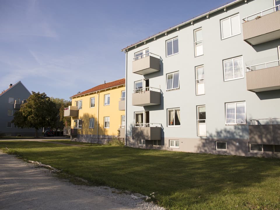 Ljusblått bostadshus framför ett gult bostadshus. Båda husen har balkonger. Framför dem är det gräsmatta. Himlen är blå.