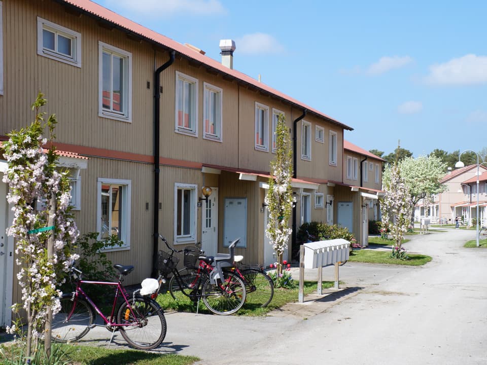 En ljusgul radhuslänga med utstickande förråd, längs med en asfalterad väg. Till varje entré går en väg och närmast i bild står tre cyklar vid entrén. Himlen är ljusblå med några moln.