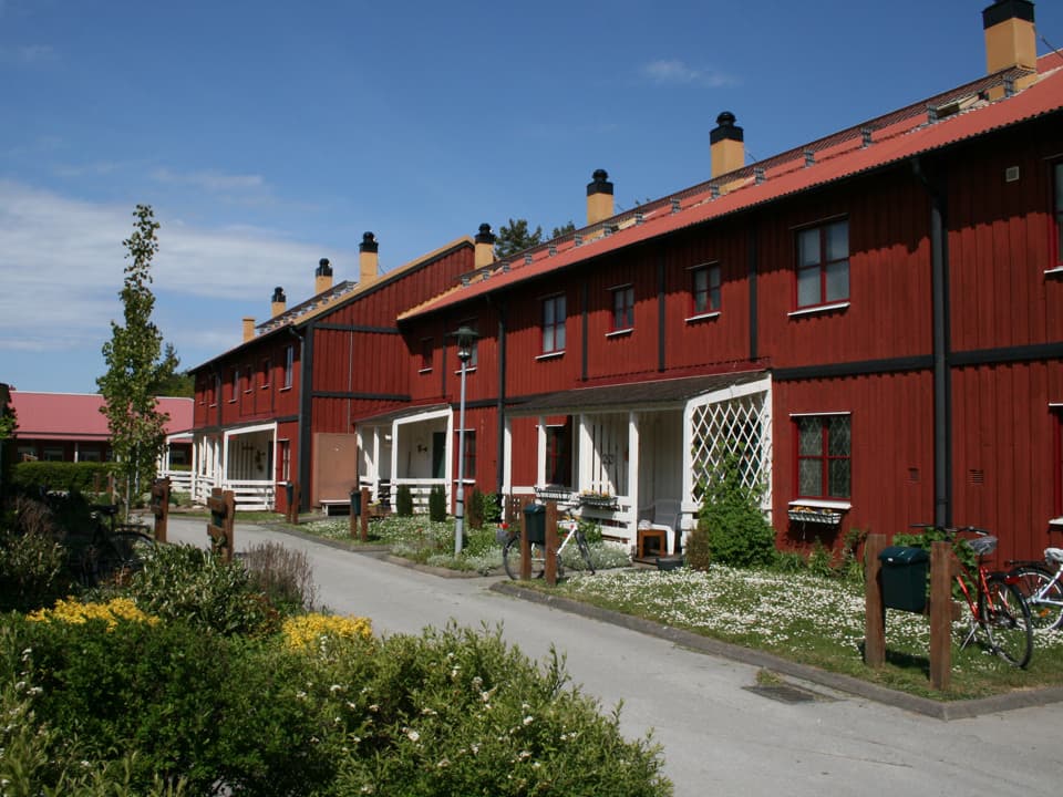 Röda radhus med vita entréer. Framför husen är en inramad gräsmatta och en asfalterad väg. Himlen är ljusblå med några moln.