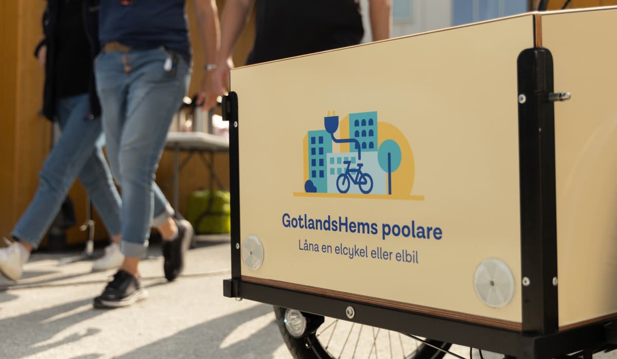 En gul lådcykel med texten "GotlandsHems poolare - Låna en elcykel eller elbil". Tvp personer med blåa jeans går förbi bredvid cykeln.