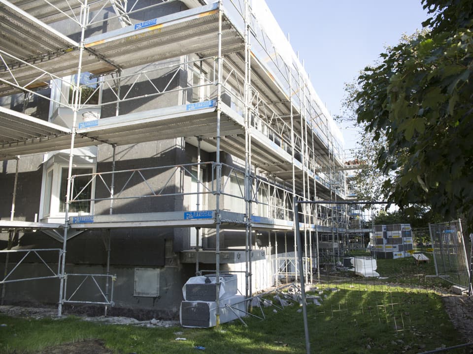 En byggställning är uppställd runt ett grått bostadshus. Det är en grön gräsmatta och ljusblå himmel.