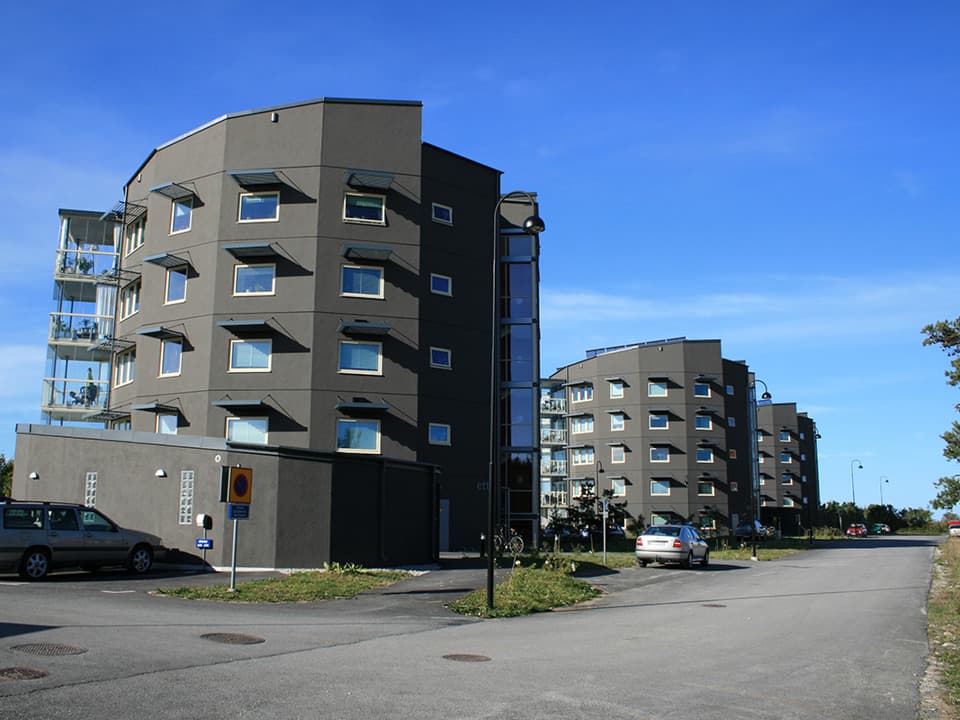 Tre svarta hexagon-formade hus på rad. Framför huset längst fram är bilar parkerade. Det går en väg förbi husen. Himlen är blå.