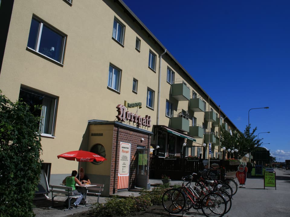 Ett avlångt ljusgult treplanshus med gröna balkonger. I bottenplan närmast kamera är entrén till konditori Norrgatt. Framför står cyklar parkerade. Himlen är blå.