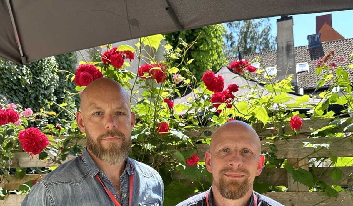 Två män med skägg och blåa skjortor ser in i kameran och ler. I bakgrunden ser man röda rosor.