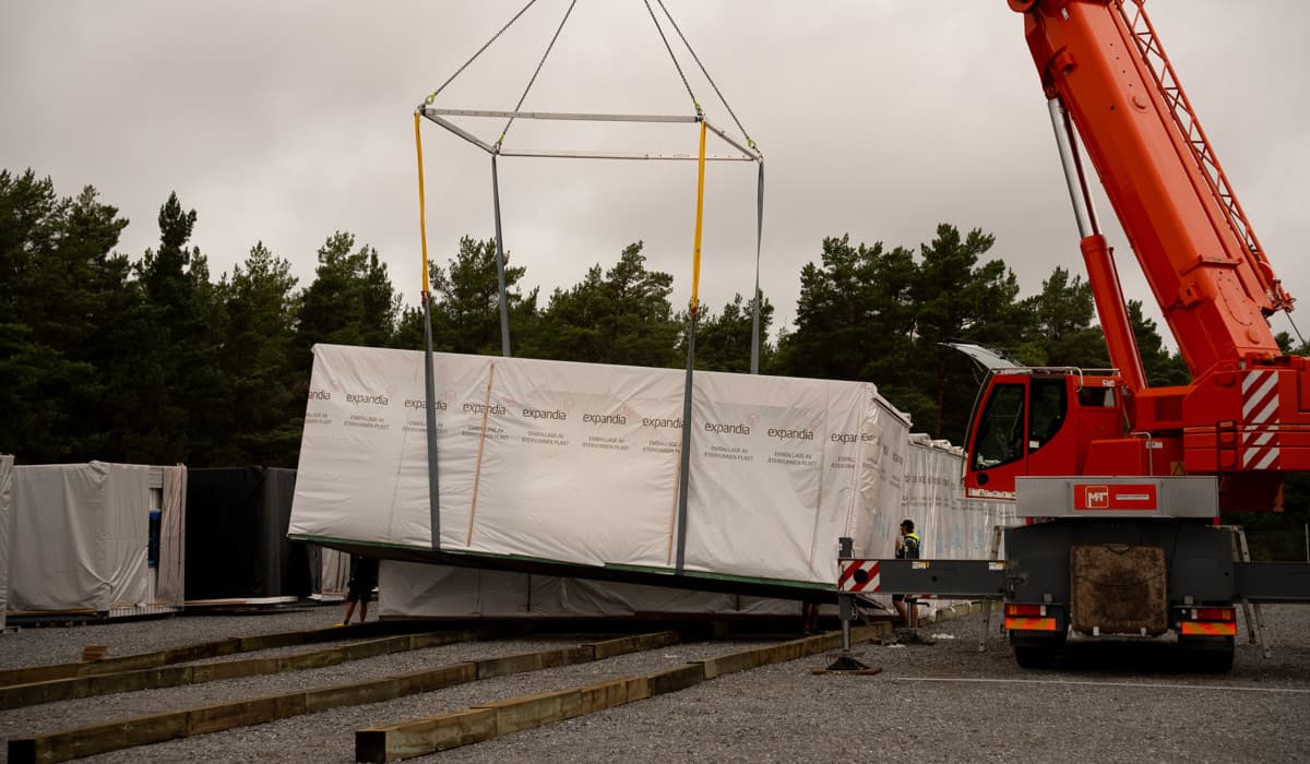 En modulbostad som ser ut som en barack är invirad i skyddsplast och lastas av på slipers. Det är en röd lyftkran som lyfter den på plats. Vädret är obestämbart mulet, gråvit himmel med gröna tallar i bakgrunder. 