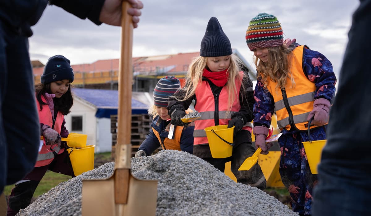 Fyra barn i femårsåldern står på en hög med grus. De har gula hinkar och spadar i händerna och är redo att gräva. I förgrunden syns en förgylld spade för vuxna, en man håller i den. 