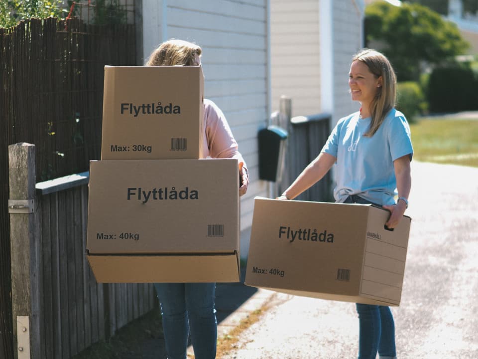 Två kvinnor bär på flyttkartonger. Den ena kvinnan har en rosa tröja och bär två kartonger, den andra kvinnan har en ljusblå t-shirt och bär en kartong. De är utomhus i ett bostadsområde.