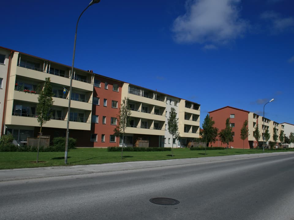 Två avlånga bostadshus med fyra plan ligger längs med en bilväg. Husen har ljusgula balkonger och fasad i rött och vitt. Det är en gräsmatta och planterade träd mellan husen och vägen. Himlen är blå med några få moln.