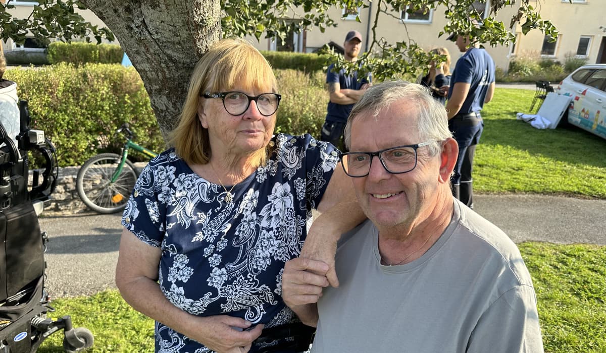 En kvinna med rött hår och svarta glasögon håller en man i handen. Mannen har grå t-shirt och ler