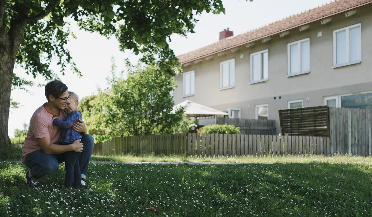 En man sitter på huk och kramar en pojke. De är utomhus under ett grönt träd på en grön gräsmatta. I bakgrunden syns ett vitt bostadshus.