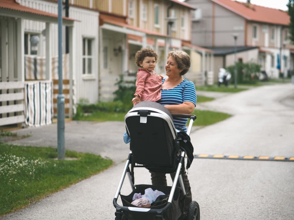 En kvinna bär en liten flicka och drar en barnvagn framför sig. De går på en väg i ett bostadsområde.