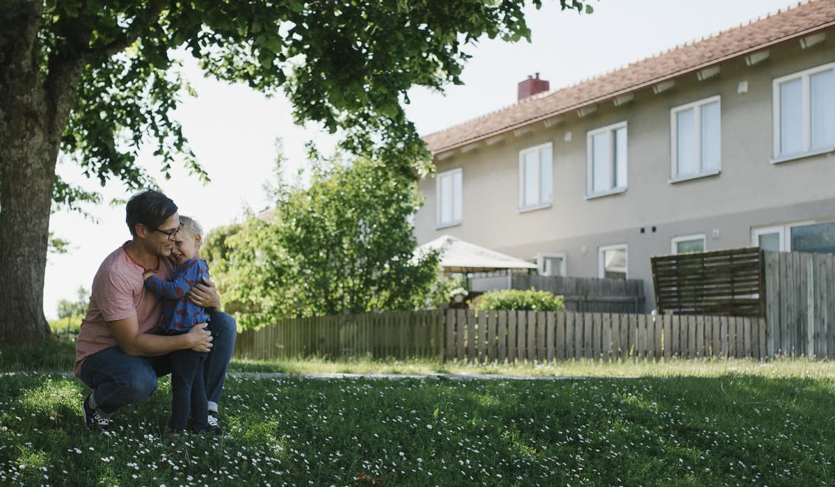 En man sitter på huk och kramar en pojke. De är utomhus under ett grönt träd på en grön gräsmatta. I bakgrunden syns ett vitt bostadshus.
