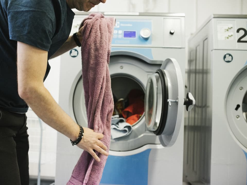 En man med svart t-shirt och svarta byxor håller i en rosa handduk. Han står i en tvättstuga framför en tvättmaskin med öppen lucka.