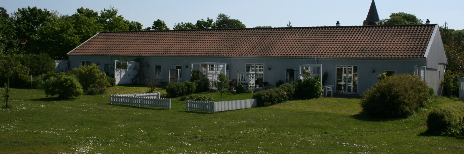 Avlångt grått hus med tegeltak. Framför huset är en gräsmatta och flera buskar. I bakgrunden syns ett kyrktorn. Himlen är ljusblå med ett moln.