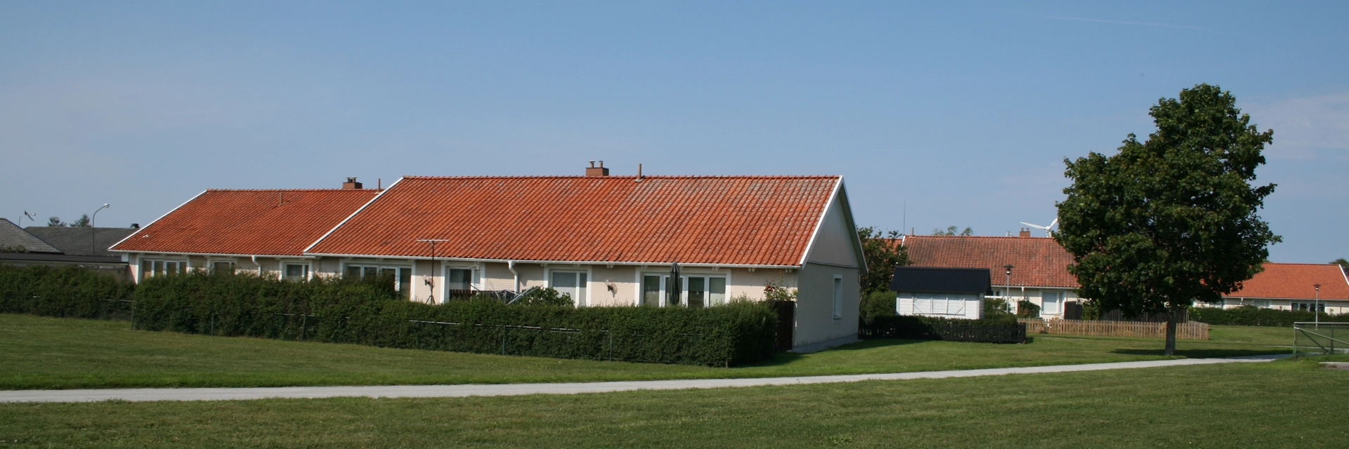Enplanshus med rosa fasad och rött tak bakom en häck. Framför är en stor gräsmatta. Himlen är ljusblå.