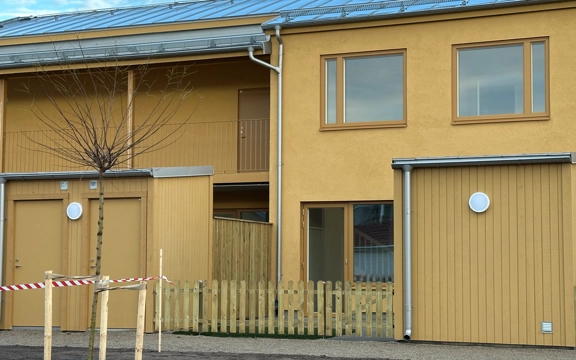 Visning av nybyggda bostäderna på Silvergatan, kvarteret Nötskrikan i Hemse. Flerfamiljshus i två våningar, gul fasad. Marken framför är bara tillplattad jord. Det är barmark men vinter. Klarblå himmel. 