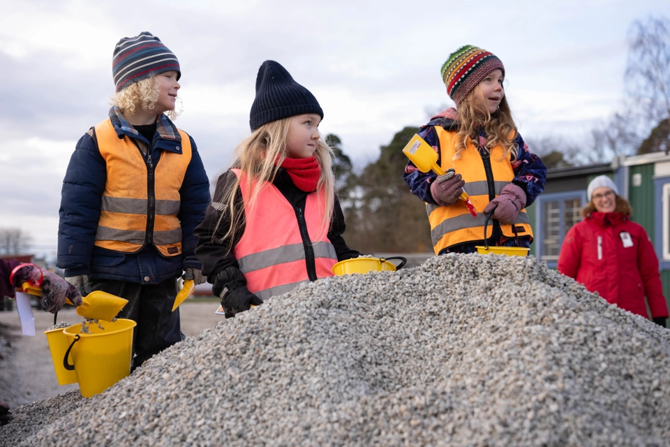 Fyra barn i femårsåldern står på en hög med grus. De har gula hinkar och spadar i händerna och är redo att gräva. I förgrunden syns en förgylld spade för vuxna, en man håller i den. 