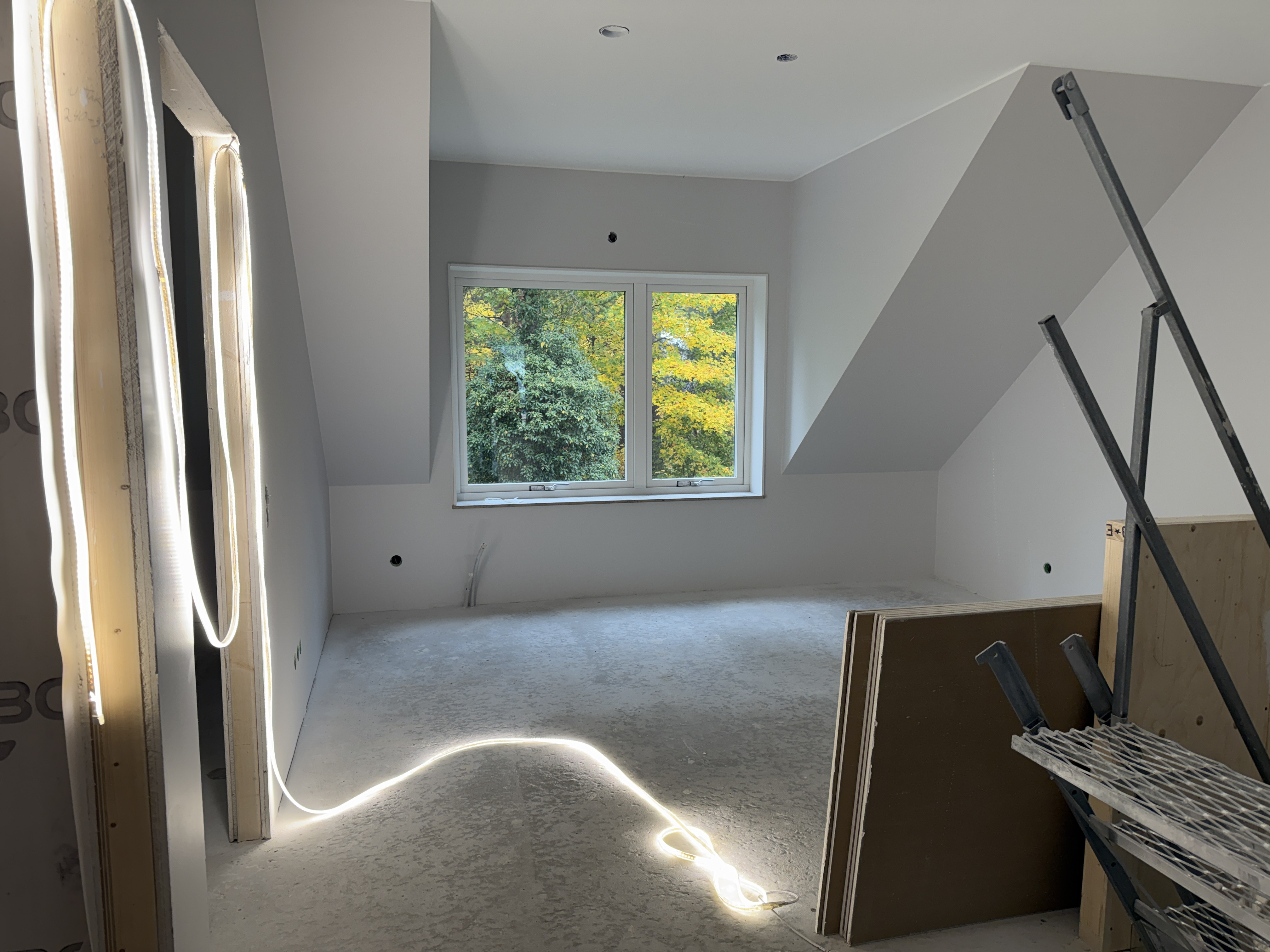 Ett tomt rum i e nybyggd bostad. Ett fönster ut om höstfärgade löv. Till höger en trappa upp till rummet.