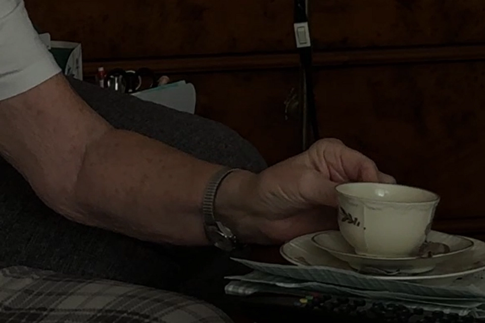 En äldre kvinna håller i en kaffekopp i ett mörkt rum.