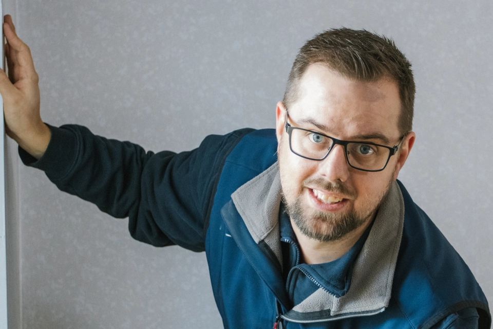 En man med blå jacka och svarta glasögon står i ett vitt rum. Han tittar in i kameran och ler. Han håller ena handen på väggen. 