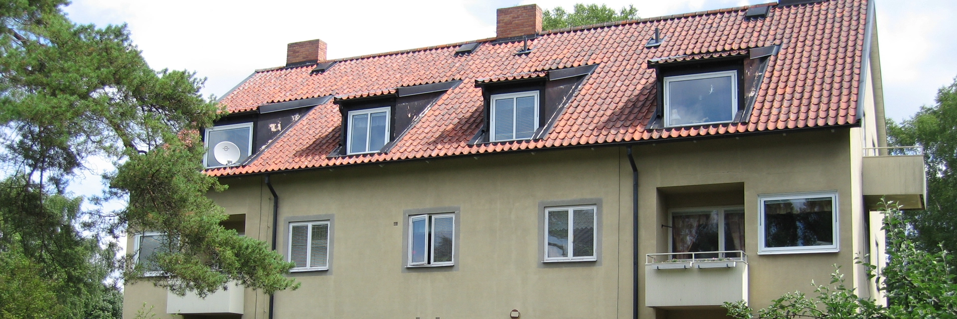 Gult bostadshus med ljusgula balkonger. Till källarplan är det vita garagedörrar, det går en smal asfalterad väg fram till huset. Runtom är det gräsmatta, buskar och träd.