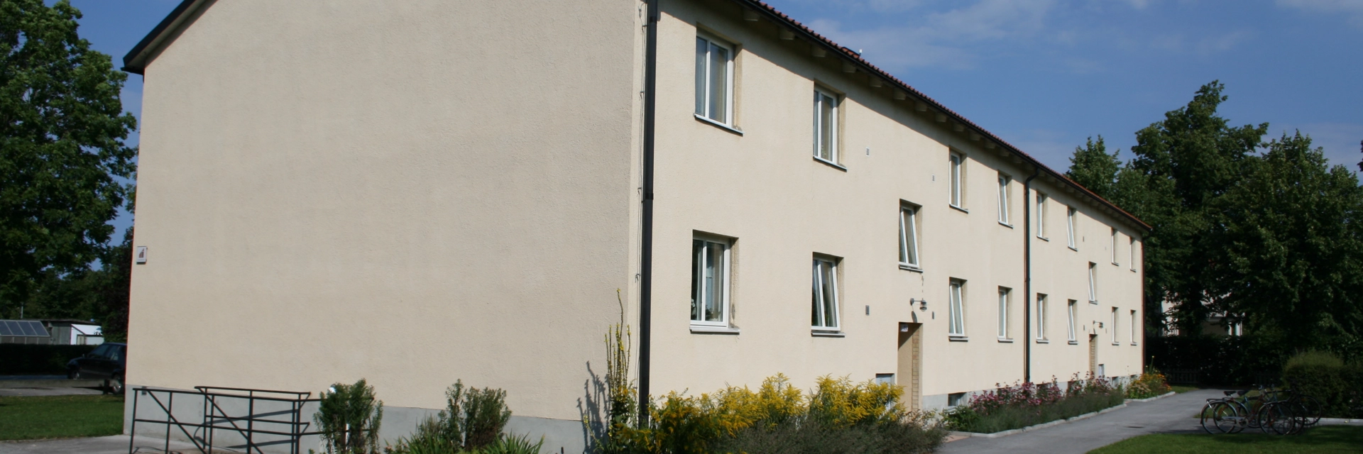 Avlångt bostadshus med beige fasad. Runt huset är en asfalterad smal väg och därefter gräsmatta.