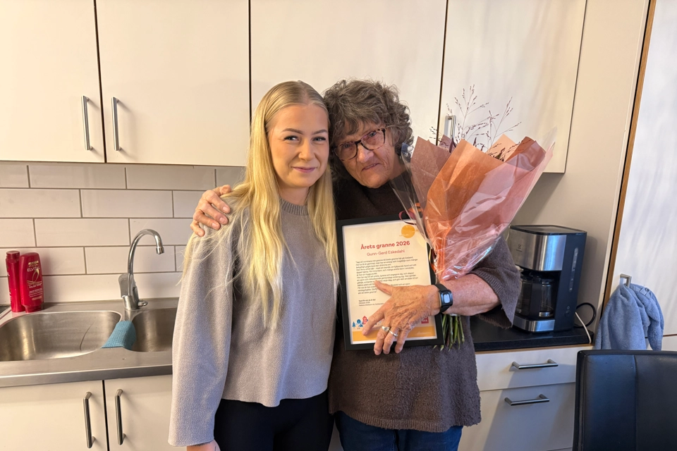 Två grannar som står håller om varandra i ett kök. 