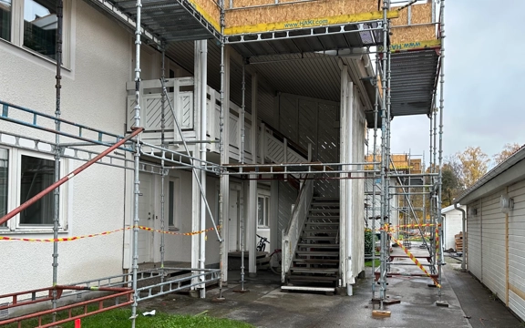 Kortsidan av ett tvåvåningshus i vitt trä är täckt av byggställningar inför en renovering. Mittemot byggnaden syns en sidobyggnad där man byter ut delar av fasaden. 