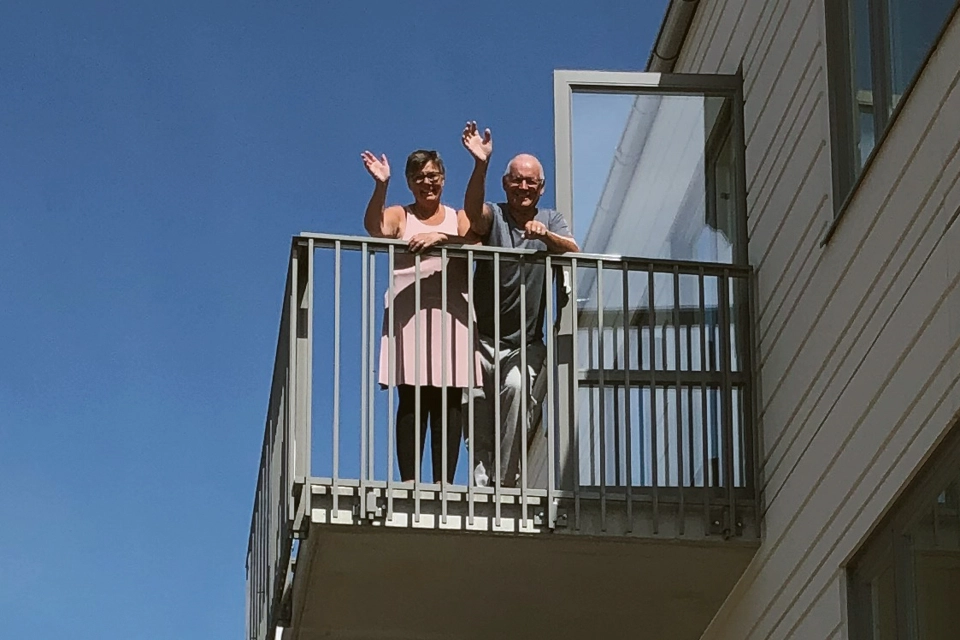 Två personer står på sin balkong och vinkar.