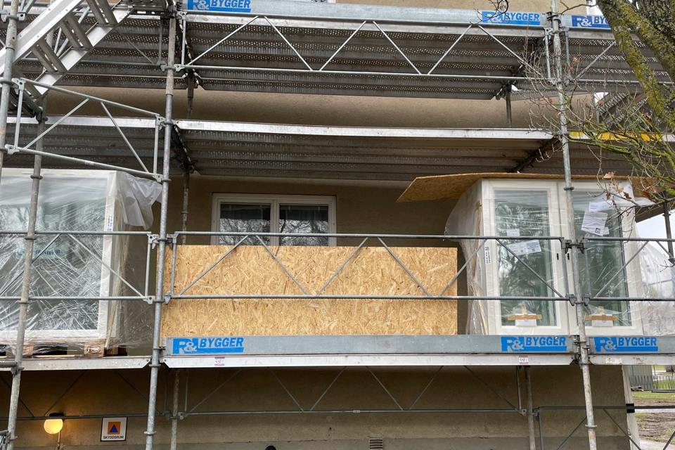 En byggställning i flera våningar på gaveln till ett beige bostadshus. Nya fönster som ska monteras står på byggställningens första våning.
