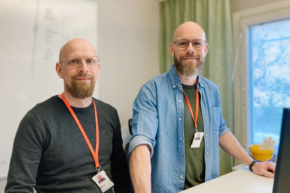 Två män med glasögon och skägg står framför en dator. De tittar in i kameran och ser glada ut.