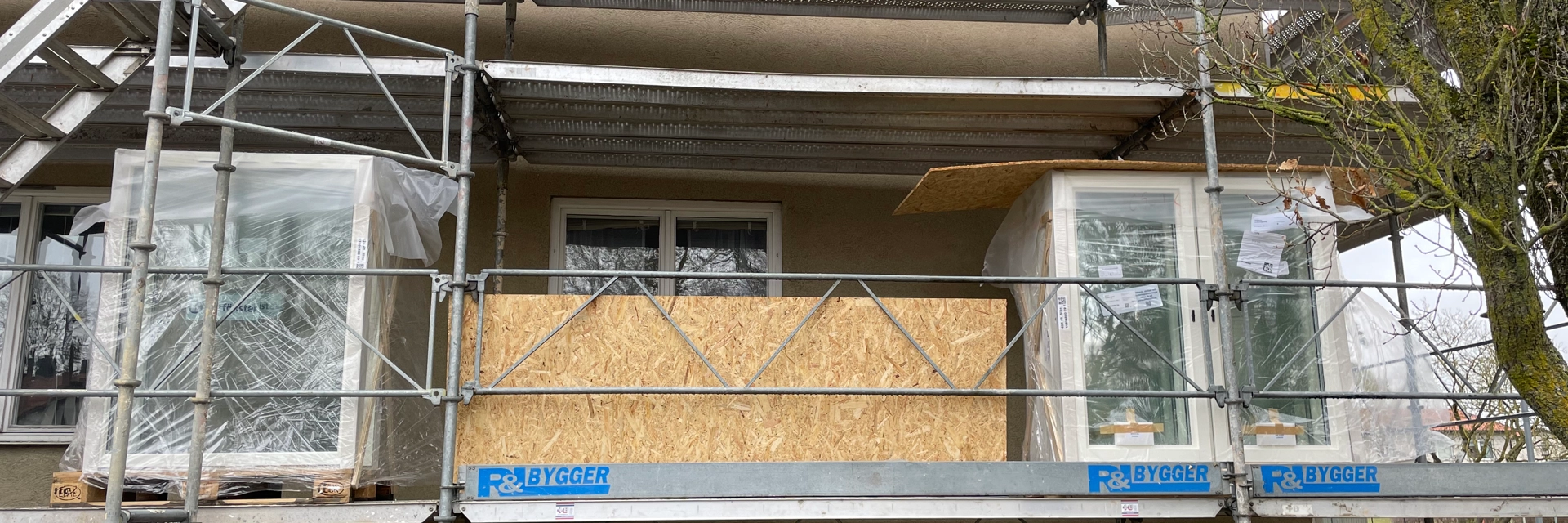 En byggställning i flera våningar på gaveln till ett beige bostadshus. Nya fönster som ska monteras står på byggställningens första våning.