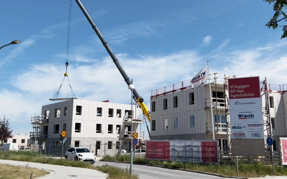 Två vita bostadshus under byggnation. En kranbil lastar material till det ena huset. Det är en byggskylt framför där det står "Vi bygger 62 nya hyresrätter" följt av företagsloggor.