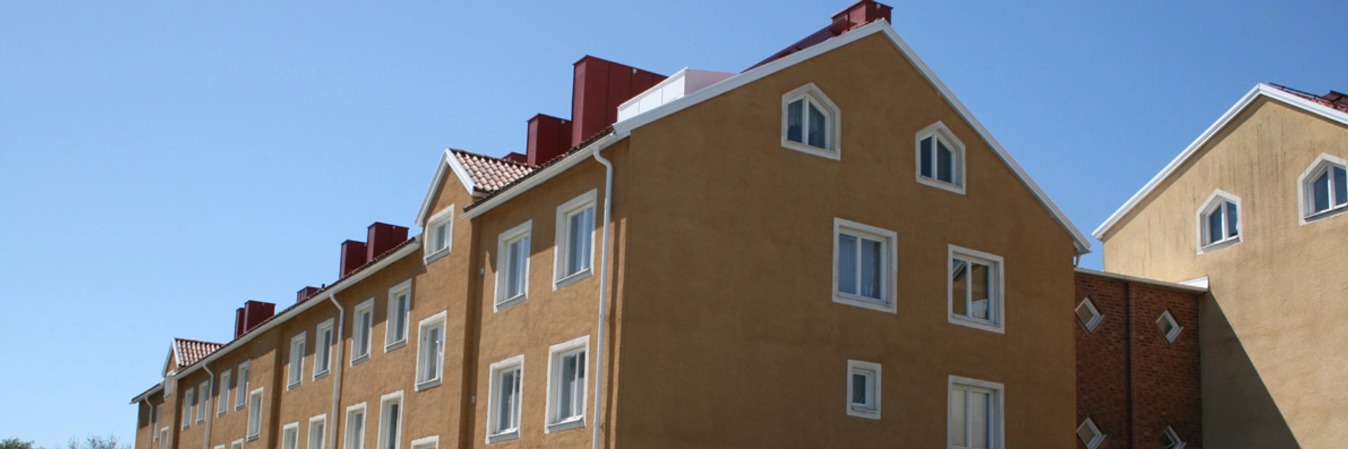 Ett gult trevåningshus med vita fönsterkarmar.Framför huset går en smal, asfalterad cykel- och gångbana och på sidan en häck.