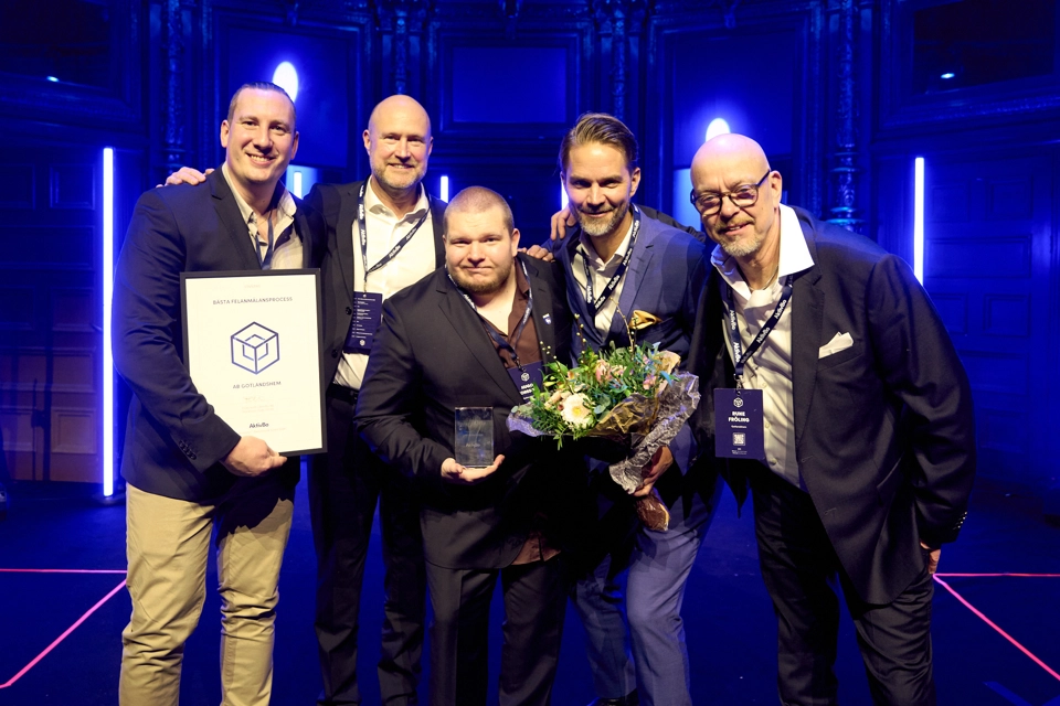 Fem män i kostym står och håller om varandra. De är glada och tittar in i kameran. En håller i ett diplom, en i ett pris och en tredje i en bukett.