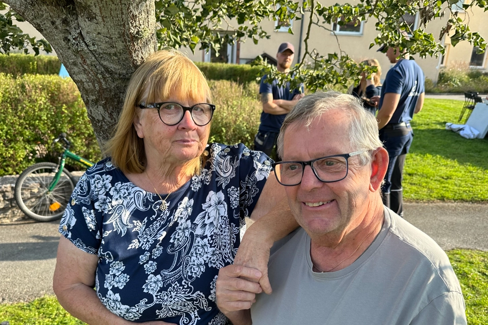 En kvinna med rött hår och svarta glasögon håller en man i handen. Mannen har grå t-shirt och ler