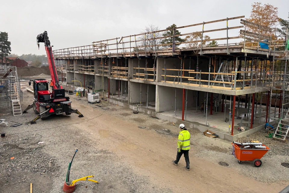 Ett husbygge där vi ser två våningar och massor av virke. En person i varselkläder går framför bygget.