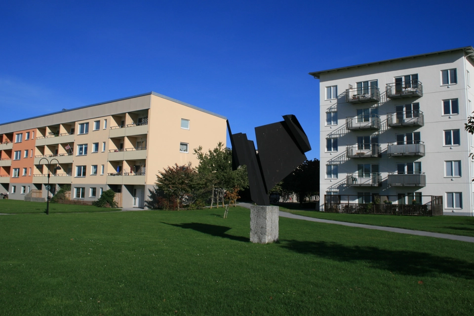 En beige byggnad och en vit byggnad står bakom en svart skulptur med abstrakt motiv.